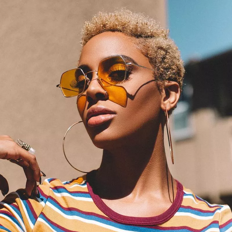 Jade Novah wears a short blonde Afro hairstyle, orange sunglasses, and large hoop earrings