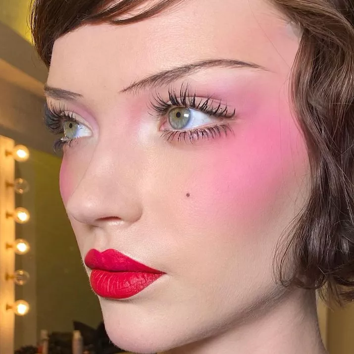 Model wears old Hollywood-inspired makeup look with bright blush, thin brows, and red lipstick