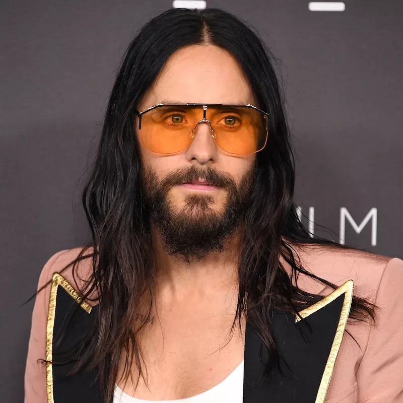 Jared Leto with long, wavy hair, orange sunglasses, and a pink blazer
