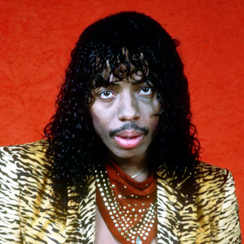 Rick James wears a wet look curly hairstyle and tiger print blazer