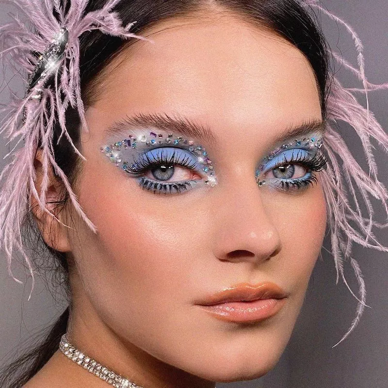 Model wears blue eyeshadow with gem graphic cut crease and pink hair feathers