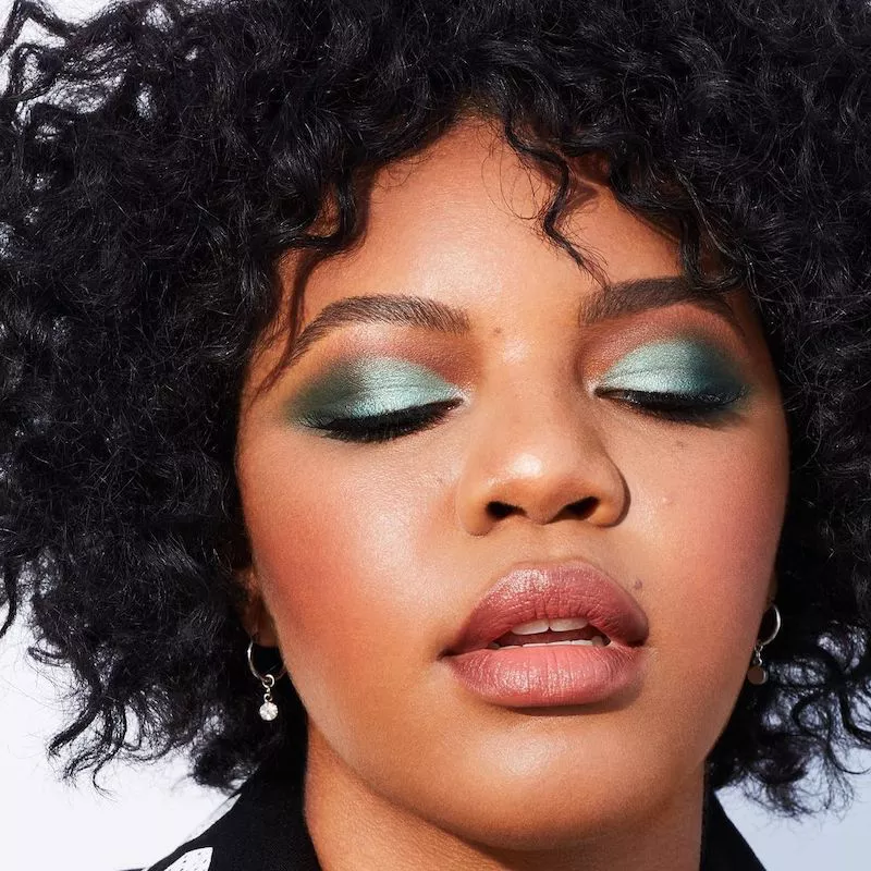 Model with green shimmery cut crease eyeshadow, peach blush, and natural curls
