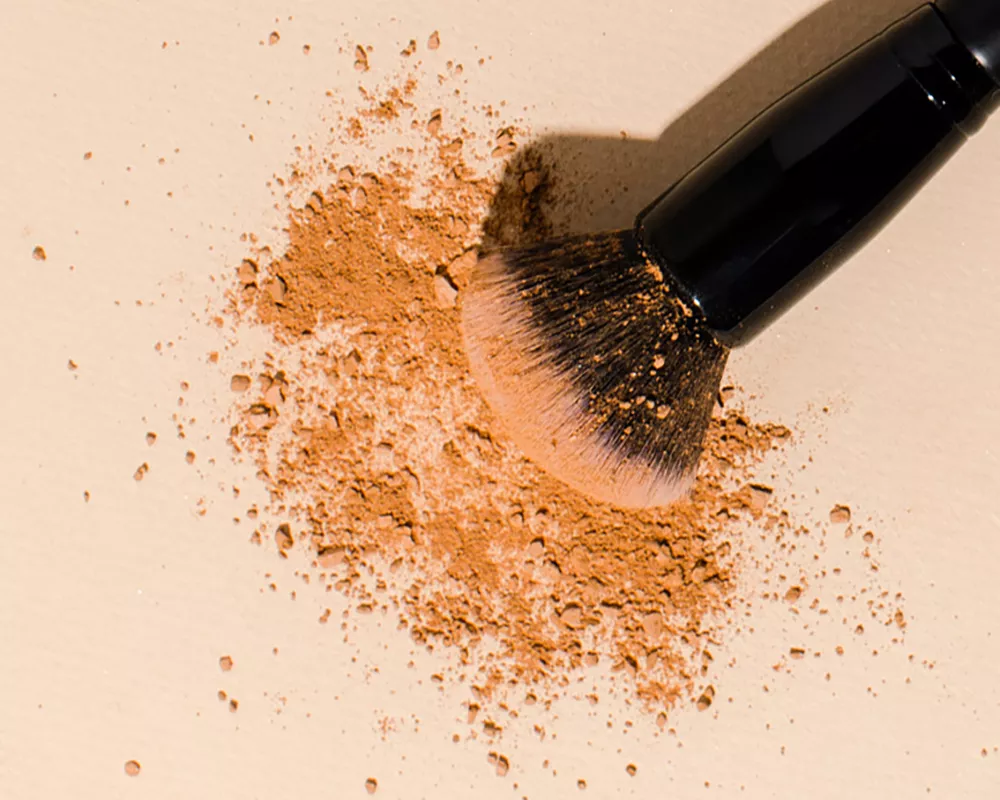 Close up of a makeup brush and powder on a cream colored background