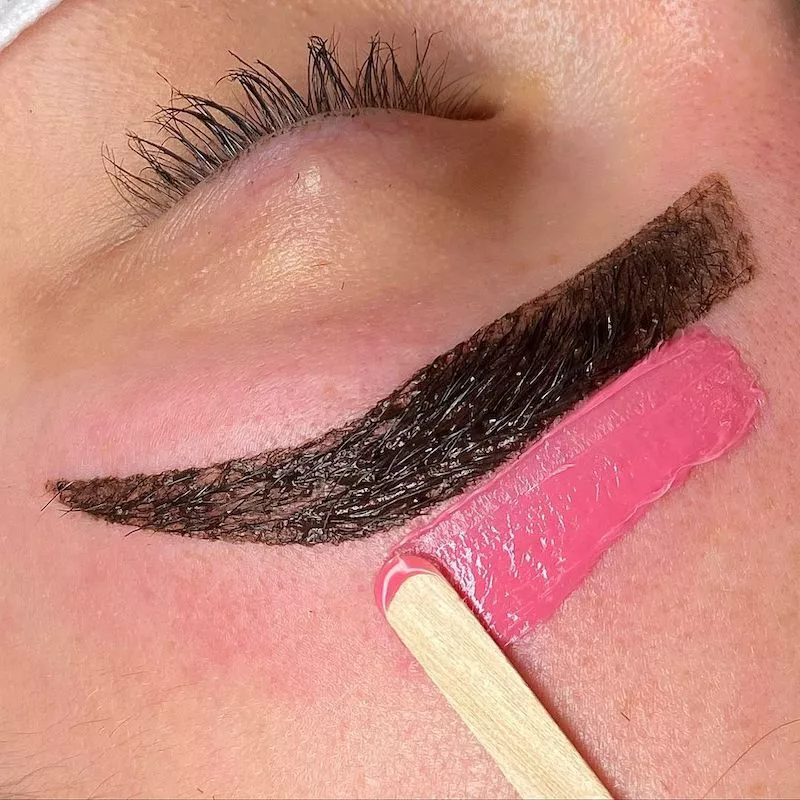 Close-up of tinted brown eyebrow with pink wax and popsicle stick nearby