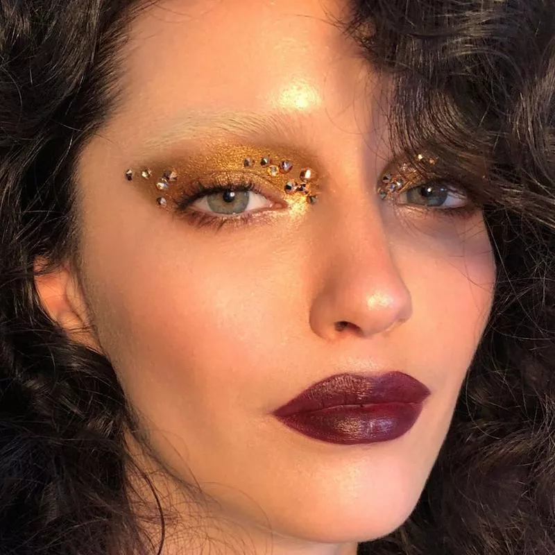 Model with burgundy lipstick, bleached eyebrows, and gold eyeshadow with gems