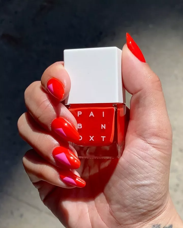 Pink and red geometric nails on a hand holding a bottle of red nail polish.