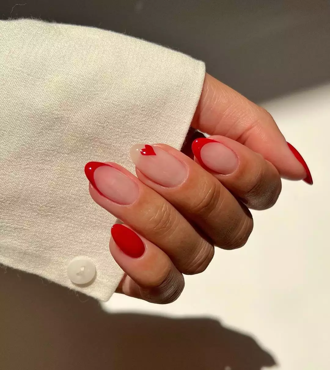 Person's hand with almond-shaped nails painted with red French tips and a tiny red heart.