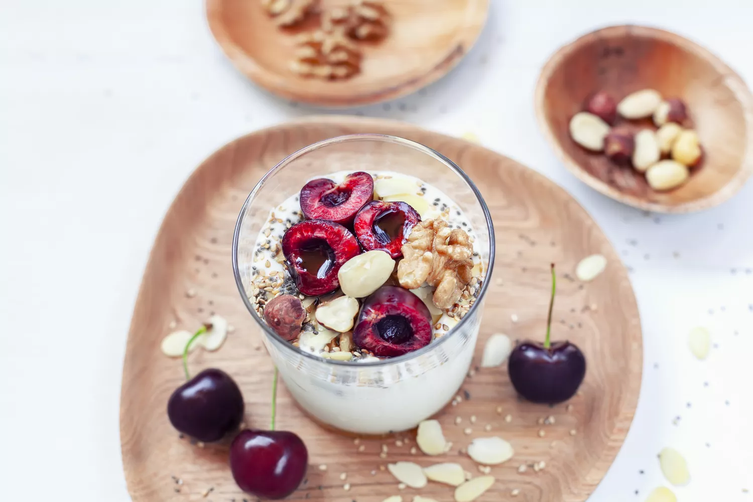 Glass of greek yogurt topped with cherries and nuts. Sitting atop a wooden plate with cherries and nuts on the side.