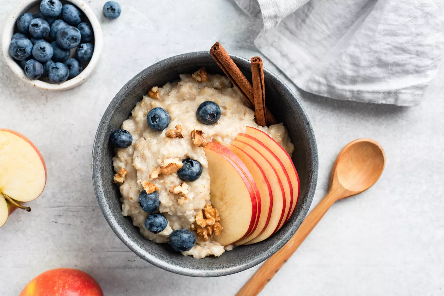 A bowl of oatmeal topped with apple, blueberries and nuts. Sitting on a counter beside a spoon, dish towel and fruit.