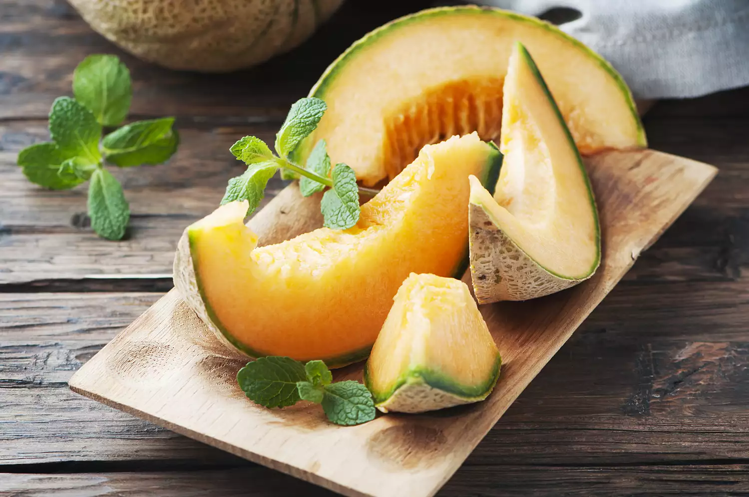 Sliced cantaloupe atop a cutting board, with a few sprigs of mint leaf/
