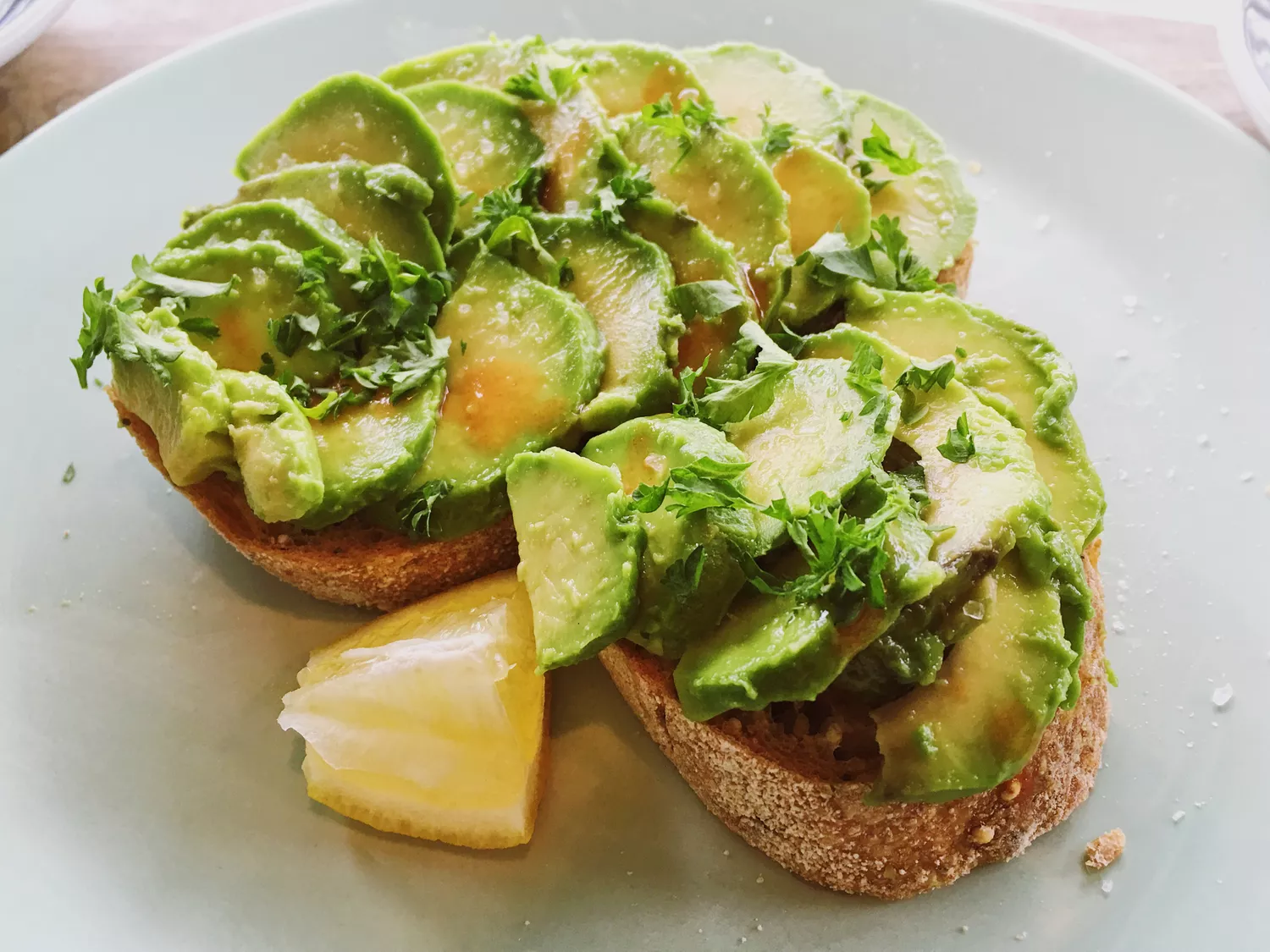 Two slices of avocado toast (two slices of toast topped with sliced avocado, herbs, oil, and salt) on a plate with a wedge of lemon beside it. 