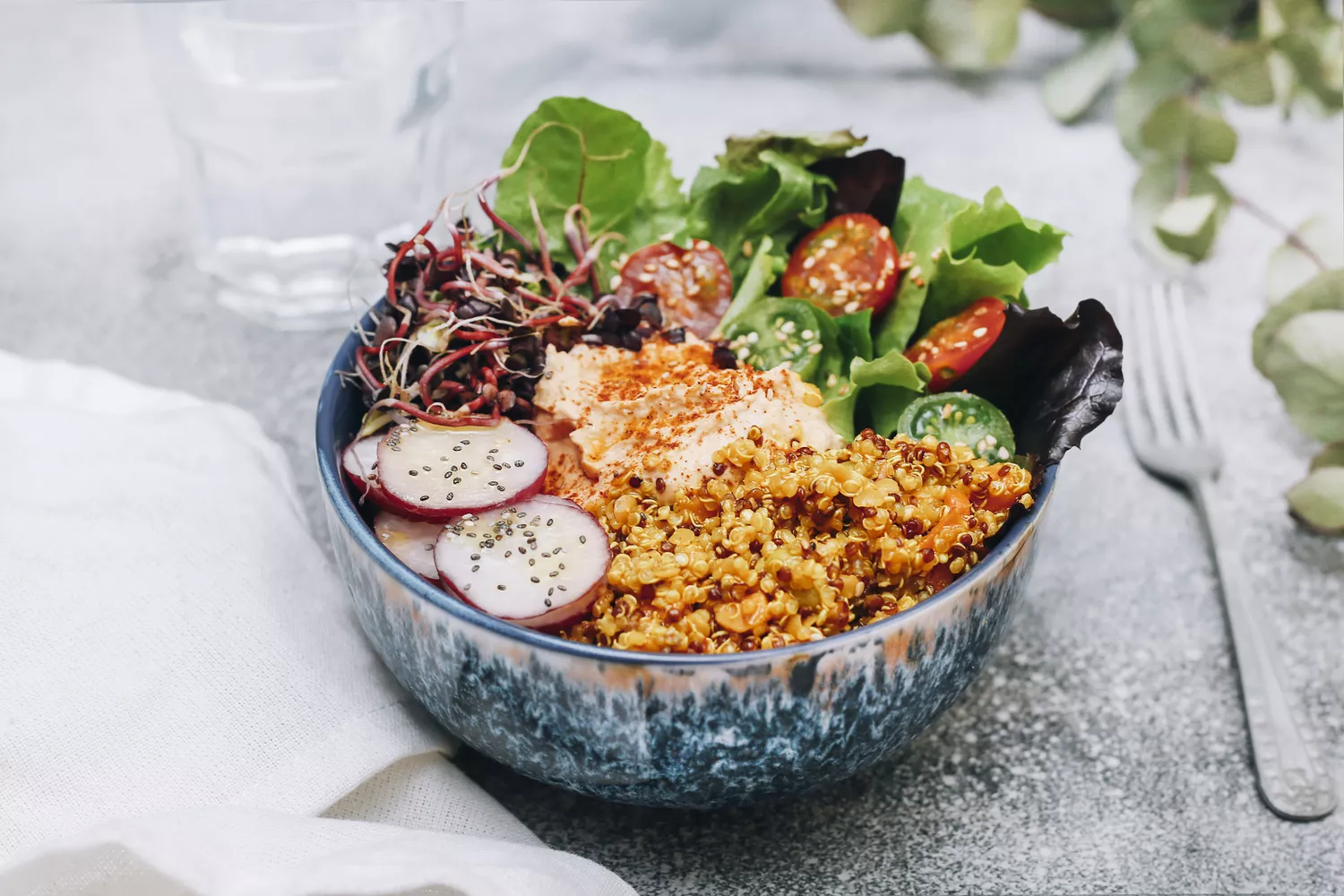 Bowl with quinoa, lettuce, radish, tomatoes and hummus. Sitting on table beside a fork.