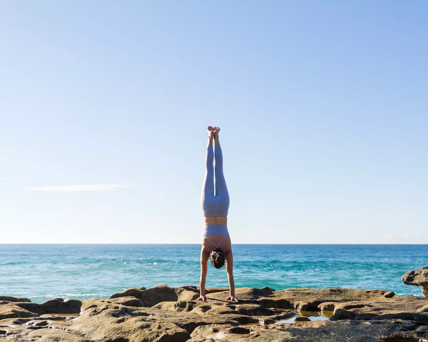 Woman in periwinkle workout set doing handstand on rocks by the sea