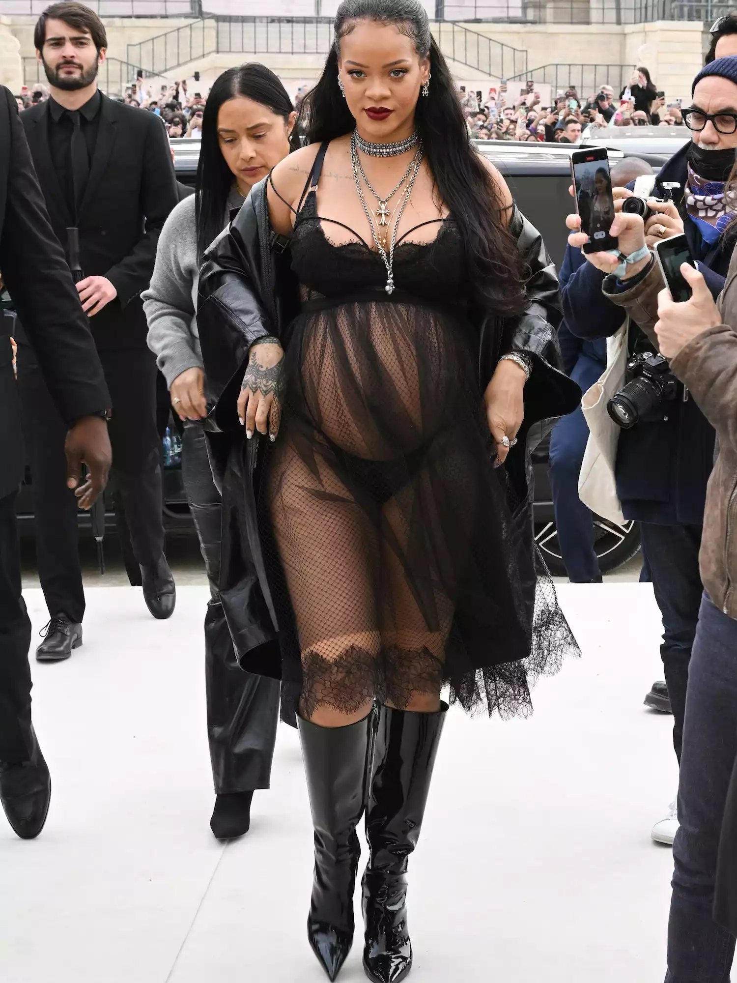 Rihanna wears a sheer black dress with lingerie, a leather trench coat, and boots to the Dior Fall 2022 show at Paris Fashion Week