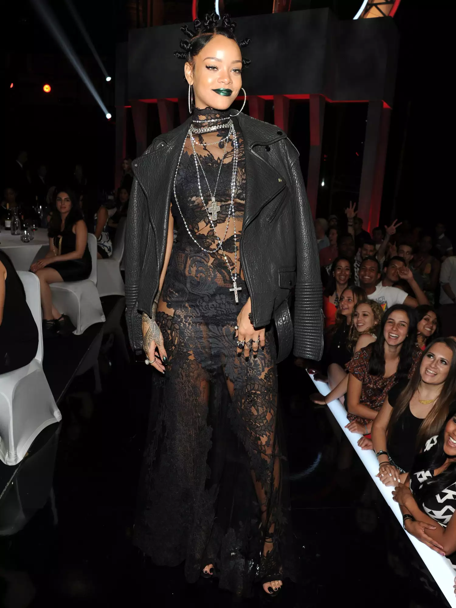 Rihanna wears a black lace dress, leather jacket, layered necklaces, hoop earrings, and bantu knots