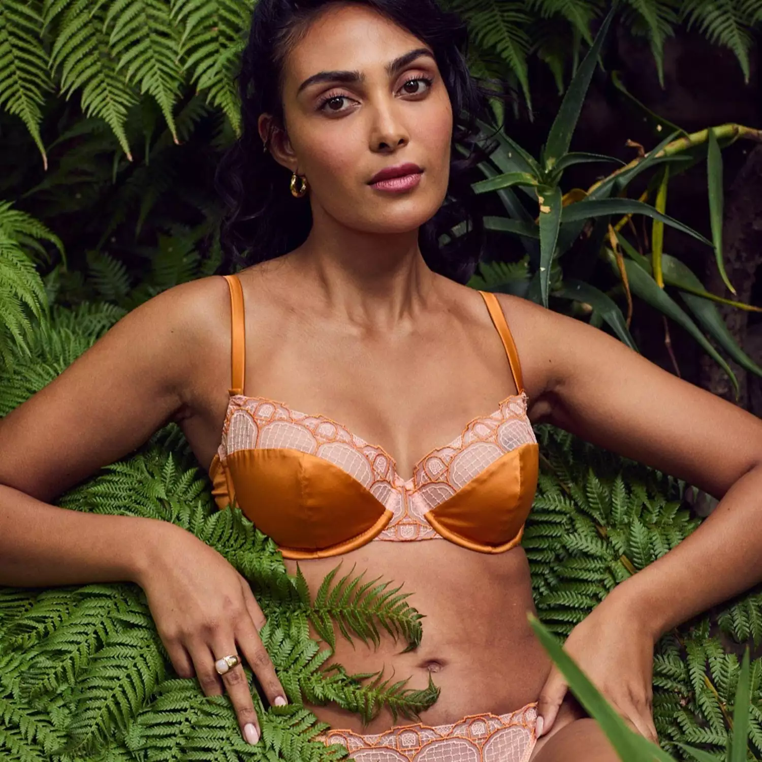 Model wearing an orange silk bra and panties set in a leafy environment