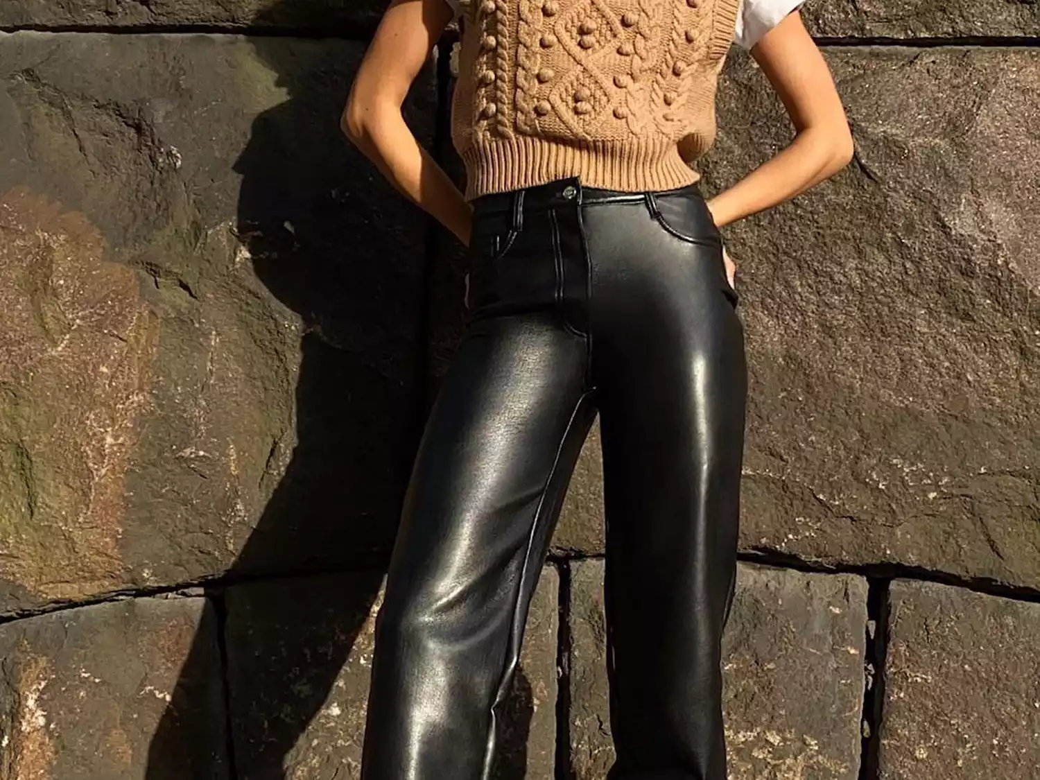Woman wears black leather pants and a taupe sweater vest against a stone wall