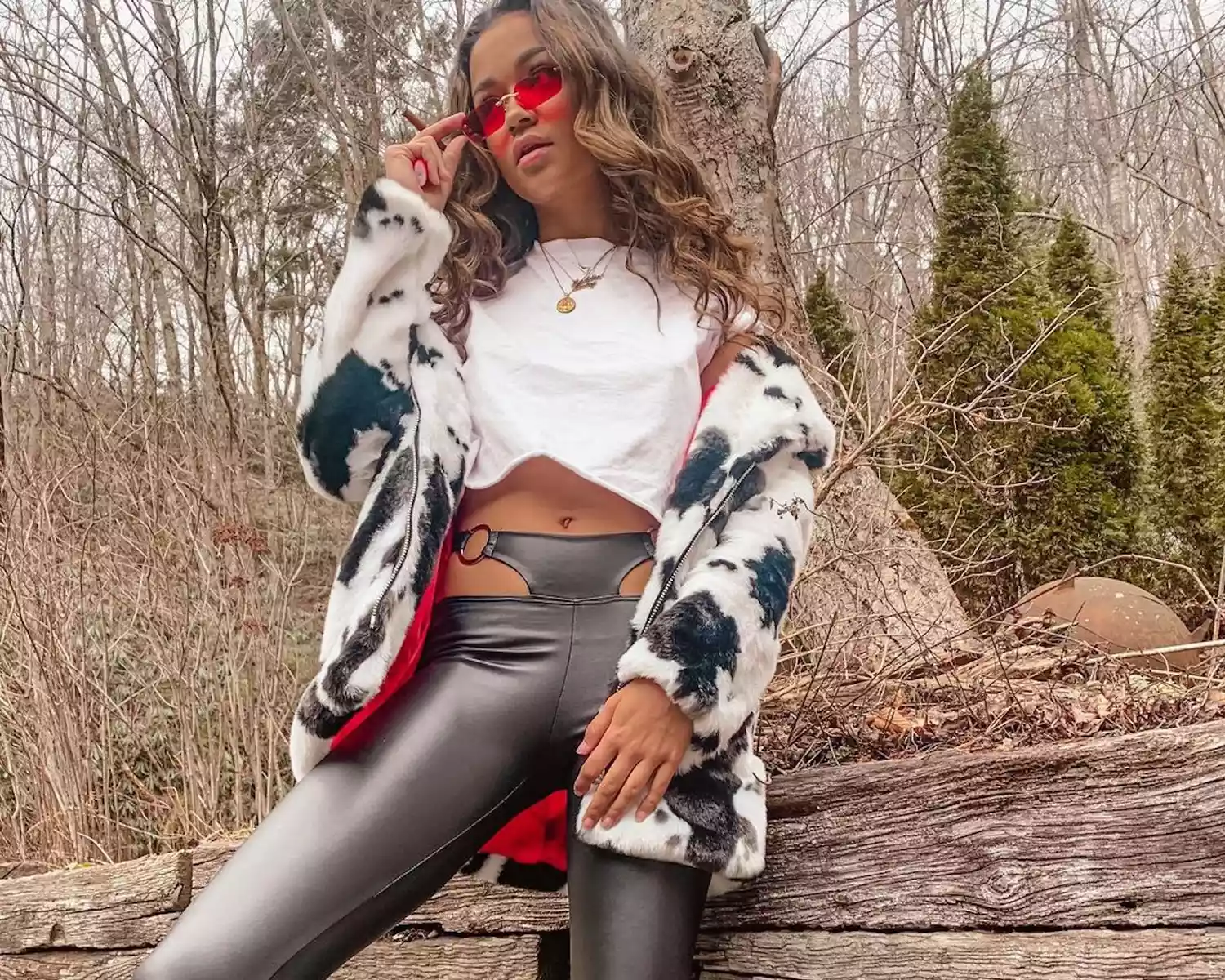 Madison Bailey wears a layered outfit with a white T-shirt, leather cutout pants, cow print faux fur jacket, and red sunglasses