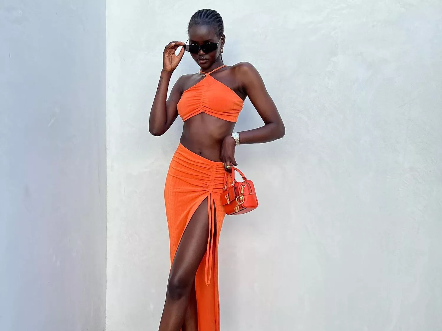 Adut Akech wears a bright orange crop top and skirt set with orange bag and black sunglasses