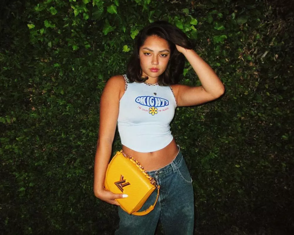 TikTok star Avani Gregg wears a white graphic tank, low-rise mom jeans, and bright yellow bag