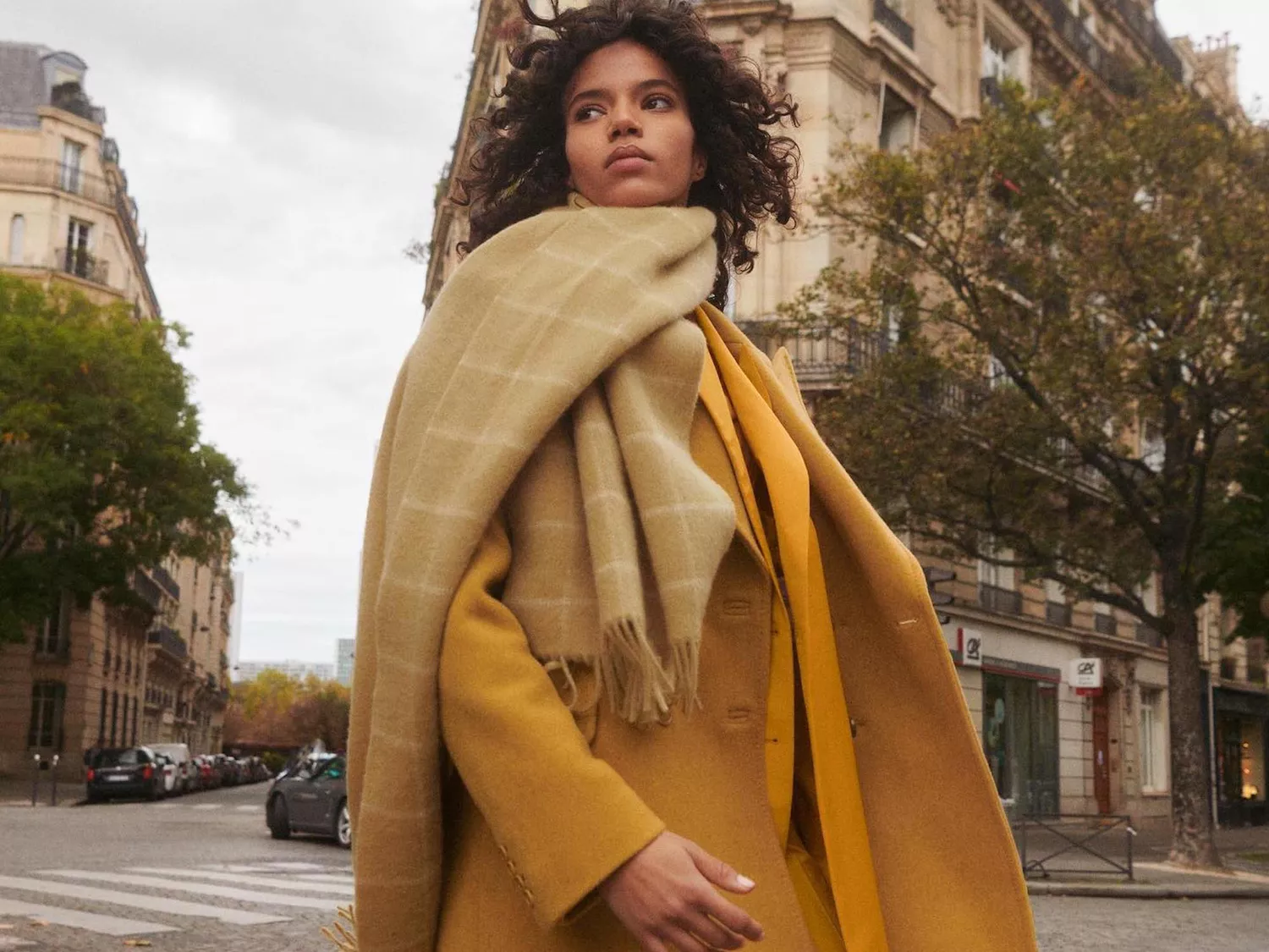 Woman with beige blanket scarf and yellow coat walking through Paris