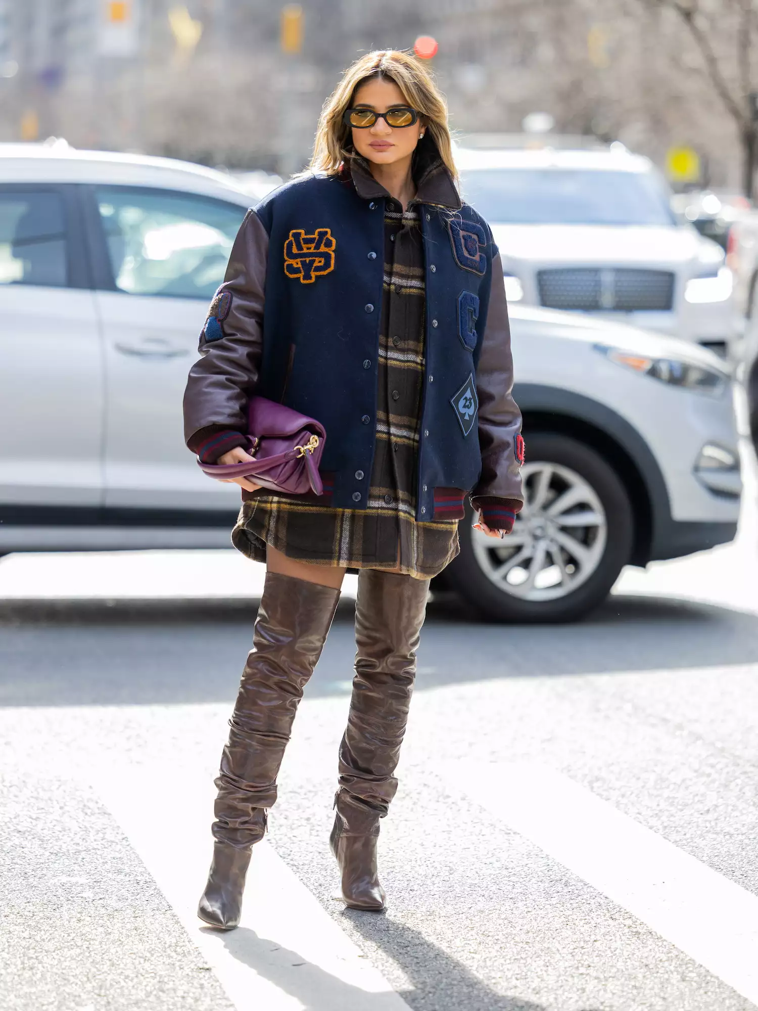 Thassia Naves wears a varsity jacket, plaid shacket, thigh-high boots, and purple bag