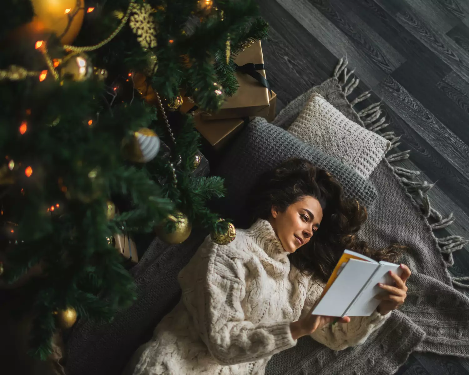 Woman reading a book while laying on pillows and blankets underneath a Christmas tree